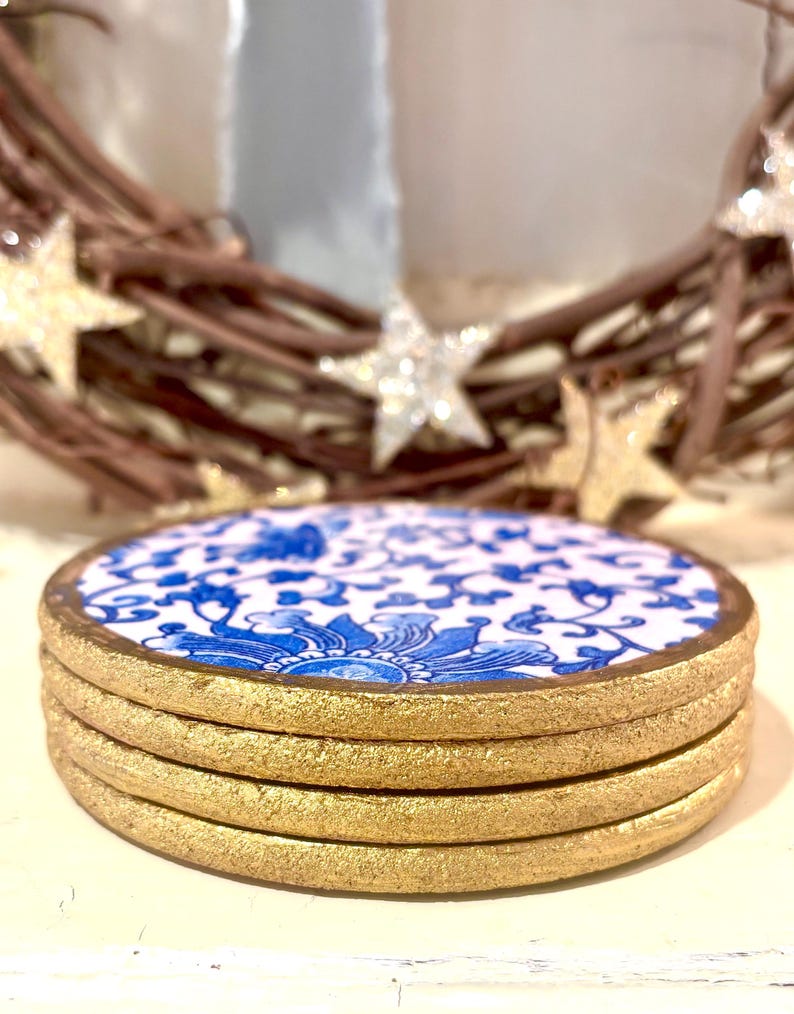 May include: A stack of four round coasters. Each coaster features a white background with a blue floral design and a gold-colored edge. The coasters are stacked on top of each other.