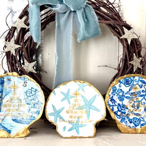 May include: A decorative wreath with a blue ribbon and three seashell ornaments. The ornaments feature blue and gold designs with inspirational text and nautical themes, including a lighthouse, starfish, and floral patterns. The wreath is made of brown twigs and adorned with glittery star accents.