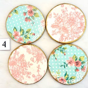 May include: Set of four round coasters with floral designs. Two coasters feature a light blue background with white polka dots and pink flowers. The other two have a white background with pink floral patterns. Each coaster has a gold painted edge and the number 4.