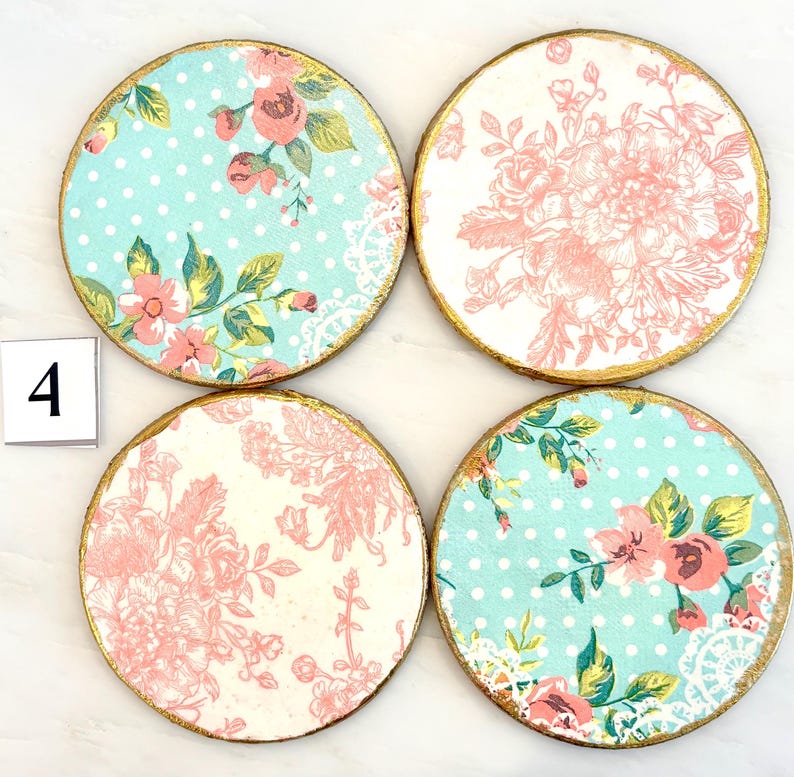 May include: Set of four round coasters with floral designs. Two coasters feature a light blue background with white polka dots and pink flowers. The other two have a white background with pink floral patterns. Each coaster has a gold painted edge and the number 4.