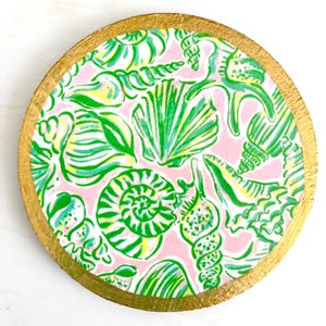K&ouml;nnte beinhalten: Runder Untersetzer mit goldfarbenem Rand. Der Untersetzer hat einen rosa Hintergrund mit einem Muster aus gr&uuml;nen, gelben und wei&szlig;en Muscheln. Das Design hat ein lebendiges K&uuml;stenmotiv.