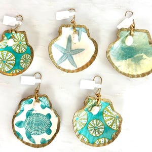 May include: Five seashell earrings with gold-painted edges and white fishhook wires. Designs include green sea urchins, a blue starfish, a coastal ocean scene, and a sea turtle. Each earring features a small white pearl bead accent and a white price tag.