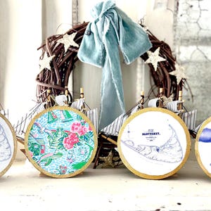 May include: Four round decorative ornaments with gold rims. Two ornaments feature a map of Nantucket, one with a floral pattern, and one with blue whales. A nautical-themed wreath and model sailboats are in the background.