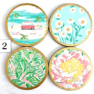 K&ouml;nnte beinhalten: Set mit vier runden Untersetzern mit goldenen R&auml;ndern. Jeder Untersetzer hat ein anderes Design: ein rosa Van auf einem Strand, wei&szlig;e G&auml;nsebl&uuml;mchen auf blauem Hintergrund, gr&uuml;ne Muscheln auf rosa und ein rosa-gelbes Blumenmuster. Die Zahl 2 ist sichtbar.