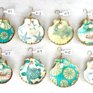 May include: Eight decorative seashell ornaments with gold-painted edges, each featuring a unique pattern including sea turtles, starfish, and tropical beach scenes. Each shell has a small white pearl accent and a white tag labeled H-1 through H-8.