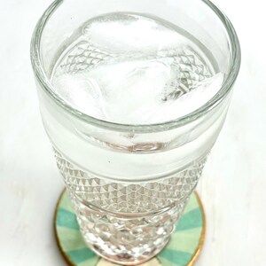 May include: A clear glass filled with water and ice cubes sits on a decorative coaster. The coaster has a green and white striped pattern with gold trim. The glass has a diamond pattern.