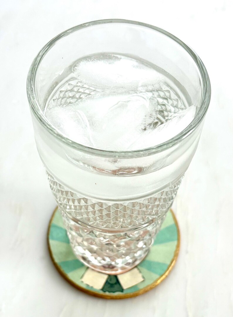 May include: A clear glass filled with water and ice cubes sits on a decorative coaster. The coaster has a green and white striped pattern with gold trim. The glass has a diamond pattern.