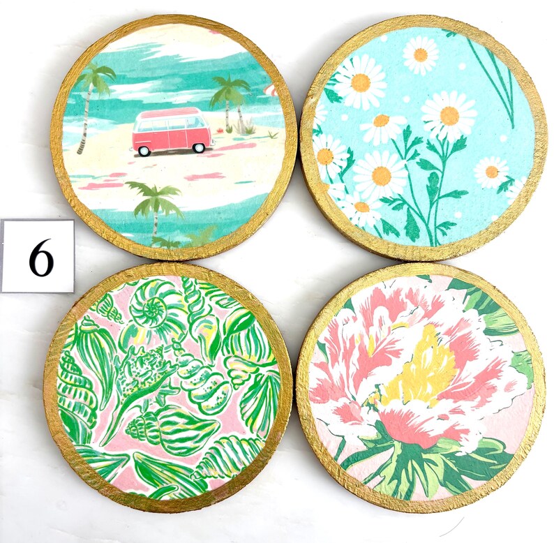 K&ouml;nnte beinhalten: Vier runde Untersetzer-Set mit goldfarbenen R&auml;ndern. Jedes Set zeigt ein anderes Design: eine Strandszene mit einem rosa Van, wei&szlig;e G&auml;nsebl&uuml;mchen auf blauem Grund, Muscheln auf rosa und ein Blumenmuster.