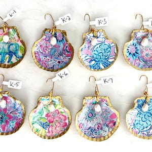 May include: Eight decorative scallop shells with gold-painted edges, each featuring a unique colorful pattern including elephants, turtles, and floral designs. Each shell has a small white pearl accent and a metal hook with a white label tag.