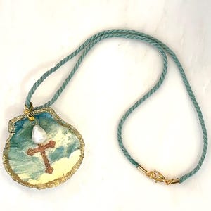 May include: A seashell pendant necklace with a cross design. The pendant features a painted scene with a cross, a pearl, and gold accents. The necklace has a teal cord with a gold clasp.