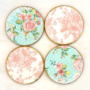 May include: Set of four round coasters with floral designs. Each coaster features a gold-painted edge. Two coasters have a light blue background with white polka dots and pink floral patterns. The other two have a white background with pink floral patterns.