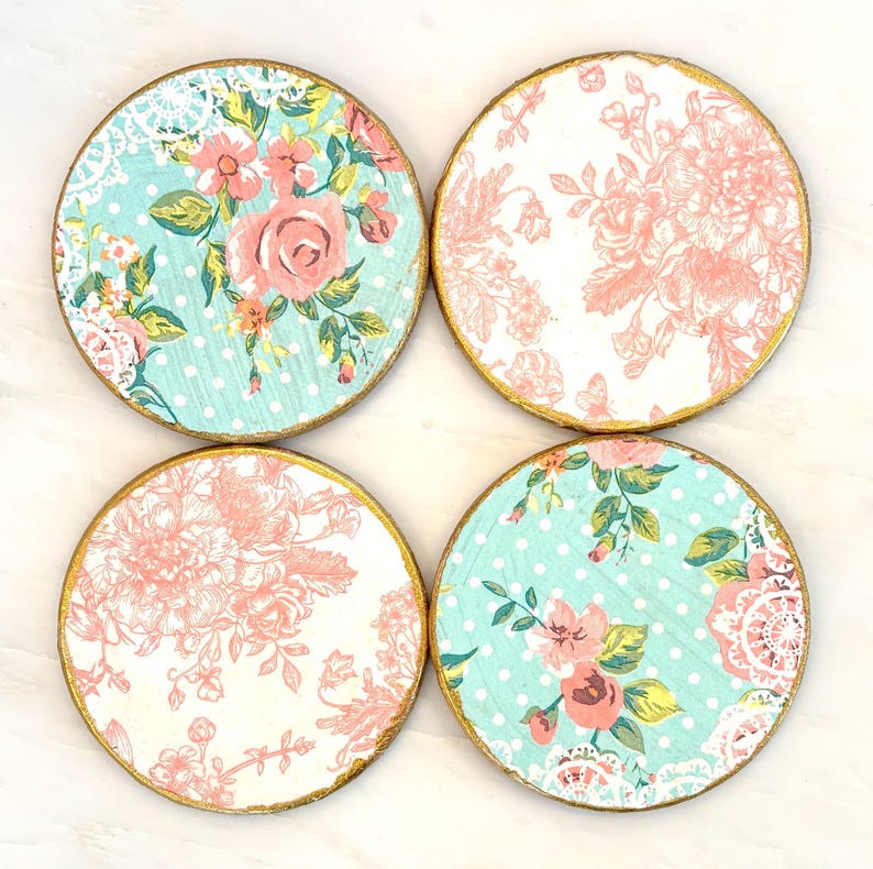 May include: Set of four round coasters with floral designs. Each coaster features a gold-painted edge. Two coasters have a light blue background with white polka dots and pink floral patterns. The other two have a white background with pink floral patterns.