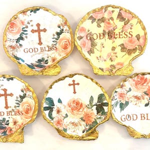 May include: Five seashell-shaped decorative plates with gold edges. Each plate features a floral design with pink and peach roses, a cross, and the words "GOD BLESS". The plates are on a white surface.