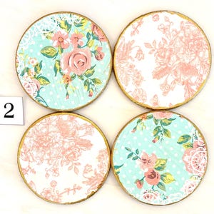May include: Four round coasters with floral designs. Two coasters feature a light blue background with white polka dots and pink roses. The other two have a white background with pink floral patterns. Each coaster has a gold edge and the number "2" in the corner.