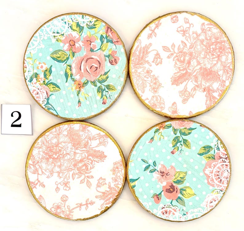 May include: Four round coasters with floral designs. Two coasters feature a light blue background with white polka dots and pink roses. The other two have a white background with pink floral patterns. Each coaster has a gold edge and the number "2" in the corner.