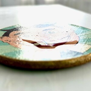 May include: A round wooden coaster with a cartoon illustration. The coaster features a gold-painted edge and a design with a face, with colors of blue, green, and peach. The coaster is a decorative item.