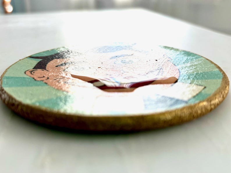 May include: A round wooden coaster with a cartoon illustration. The coaster features a gold-painted edge and a design with a face, with colors of blue, green, and peach. The coaster is a decorative item.
