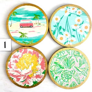 K&ouml;nnte beinhalten: Set aus vier runden Untersetzern mit goldenen R&auml;ndern. Jedes Set zeigt ein anderes Design: eine Strandszene mit einem roten Van, wei&szlig;e G&auml;nsebl&uuml;mchen auf blauem Hintergrund, ein rosa-gelbes Blumenmuster und ein gr&uuml;nes Muschelmuster.