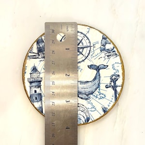 K&ouml;nnte beinhalten: Ein runder Untersetzer mit nautischem Thema, mit einem Wal, Leuchtturm, Kompass und Karte in Blau und Wei&szlig;. Der Untersetzer hat einen goldfarbenen Rand und misst etwa 10 cm im Durchmesser.