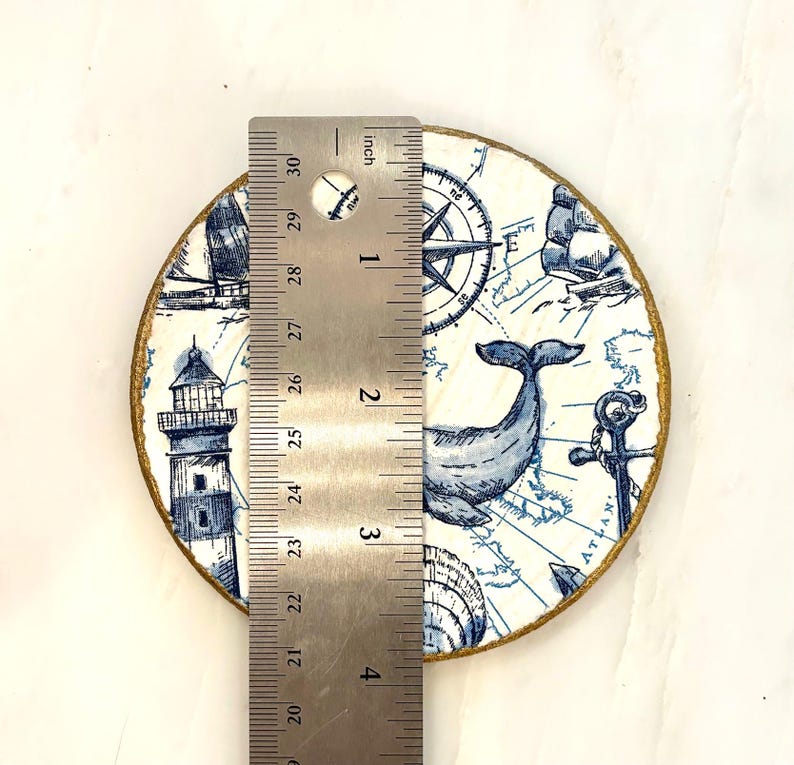 K&ouml;nnte beinhalten: Ein runder Untersetzer mit nautischem Thema, mit einem Wal, Leuchtturm, Kompass und Karte in Blau und Wei&szlig;. Der Untersetzer hat einen goldfarbenen Rand und misst etwa 10 cm im Durchmesser.
