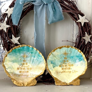 May include: Two seashell-shaped decorative dishes with gold trim and painted interiors. The dishes have a blue and yellow painted design with gold text that reads "Look for Christ and you will find Him and with Him Everything Else." A brown wreath with silver stars and a blue ribbon are in the background.