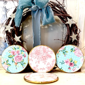 May include: A decorative wreath with a blue velvet bow and glittery star accents. Three round coasters with floral patterns in pink and blue are displayed in front of the wreath. The coasters have gold edges.