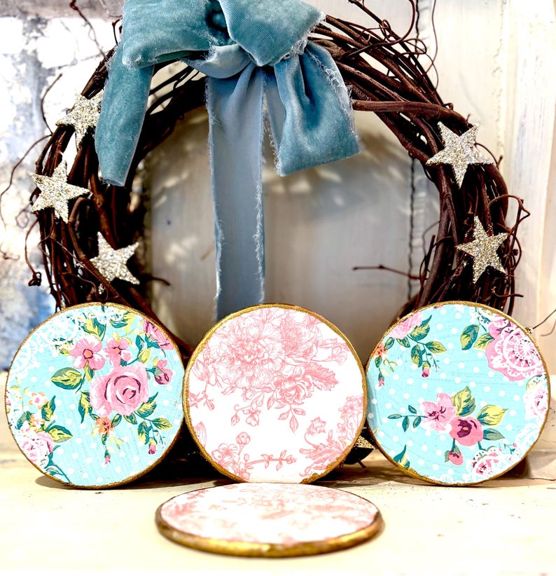 May include: A decorative wreath with a blue velvet bow and glittery star accents. Three round coasters with floral patterns in pink and blue are displayed in front of the wreath. The coasters have gold edges.