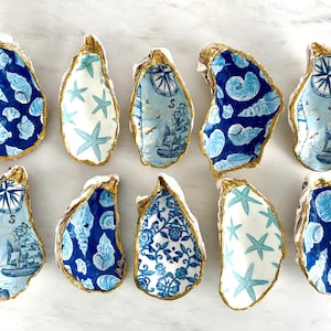 May include: A collection of hand-painted oyster shells with nautical designs. Each shell features a gold-painted edge and is decorated with blue and white patterns, including seashells, starfish, and sailing ships. These decorative shells are perfect for coastal decor.