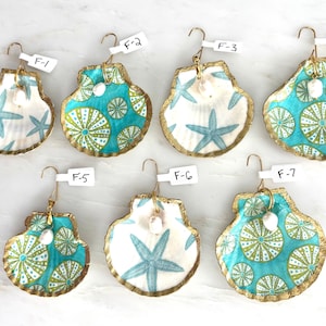 May include: Seven decorative scallop shells with gold-painted edges, each featuring a metal hook and a small white pearl-like bead. The shells display patterns of light blue starfish or green and teal sea urchins, labeled F-1 through F-7.