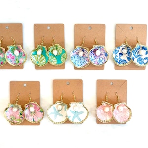May include: A collection of seashell-shaped earrings on brown display cards. The earrings feature various floral and patterned fabrics in shades of pink, blue, and green, with gold trim and pearl accents. The earrings are designed with gold-colored hooks.