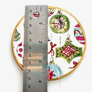 May include: A round, decorative Christmas ornament with a gold edge. The ornament features a white background with colorful, hand-painted Christmas ornament designs. The ornament is approximately 4 inches in diameter.