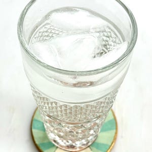 K&ouml;nnte beinhalten: Ein klares Glas gef&uuml;llt mit Wasser und Eisw&uuml;rfeln, das auf einem runden Untersetzer steht. Der Untersetzer hat ein gr&uuml;n-blaues Streifenmuster mit einem goldenen Rand. Das Glas hat ein Rautenmuster.