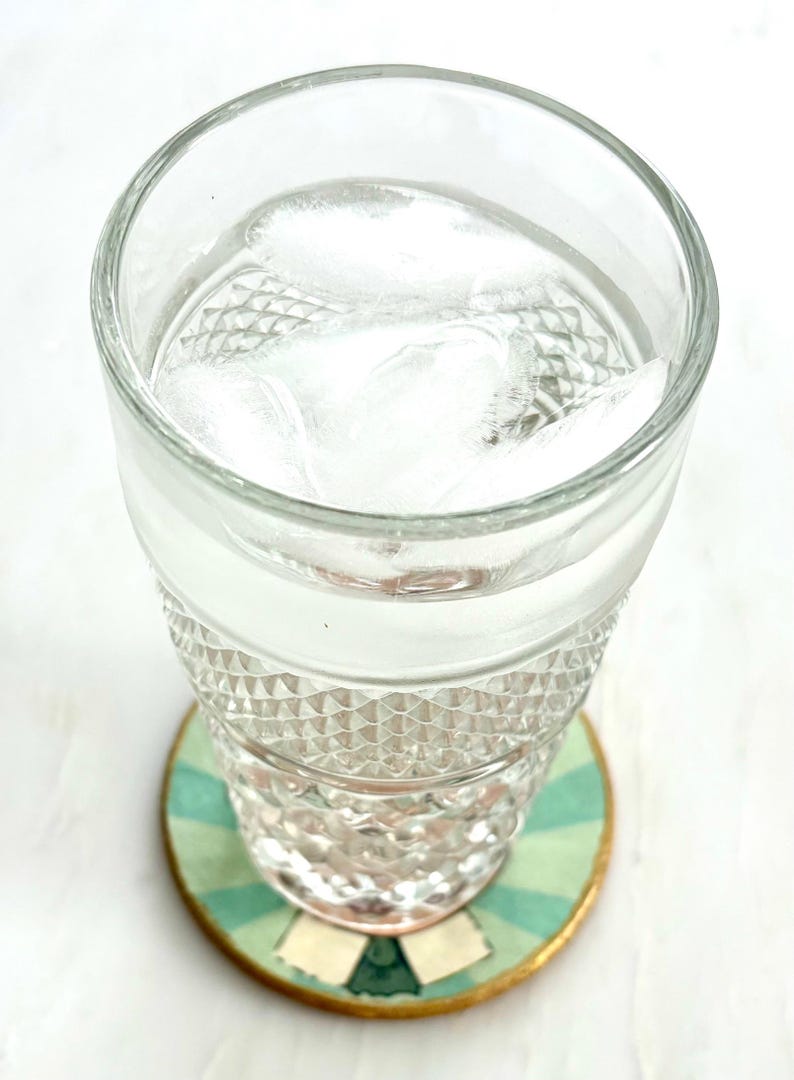 K&ouml;nnte beinhalten: Ein klares Glas gef&uuml;llt mit Wasser und Eisw&uuml;rfeln, das auf einem runden Untersetzer steht. Der Untersetzer hat ein gr&uuml;n-blaues Streifenmuster mit einem goldenen Rand. Das Glas hat ein Rautenmuster.