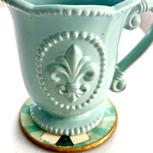 May include: A light teal ceramic mug with a fleur-de-lis emblem, set on a round coaster. The mug has a decorative handle and a scalloped rim. The coaster features a green and white striped pattern with a gold border.