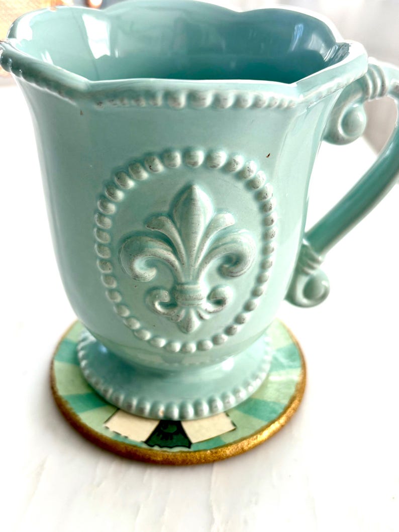 May include: A light teal ceramic mug with a fleur-de-lis emblem, set on a round coaster. The mug has a decorative handle and a scalloped rim. The coaster features a green and white striped pattern with a gold border.