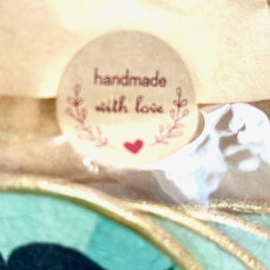 May include: A round, light brown sticker with the words "handmade with love" in a burgundy script. The sticker has a floral design and a small red heart. The sticker is on a light brown surface.