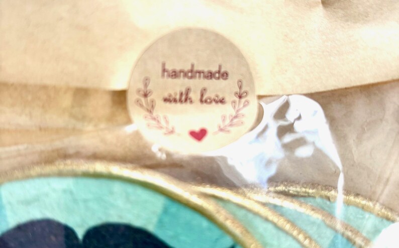 May include: A round, light brown sticker with the words "handmade with love" in a burgundy script. The sticker has a floral design and a small red heart. The sticker is on a light brown surface.