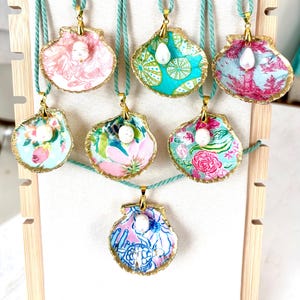 May include: A display of seven seashell pendants with gold trim, each featuring a unique fabric pattern in shades of pink, blue, and green. Each pendant is strung on a teal cord and accented with a pearl. The pendants are displayed on a wooden stand.