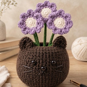 May include: A dark brown crocheted cat-shaped planter featuring two small ears, black bead eyes, a black bead nose, and embroidered whiskers. Three crocheted flowers with light purple petals and cream-coloured centres on green stems are placed inside.