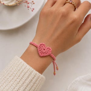 May include: A pink crochet bracelet featuring a heart-shaped charm. The bracelet is made of pink yarn and has a sliding knot closure with gold-colored beads. The bracelet is worn on a wrist.