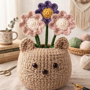 May include: A tan crocheted cat-shaped planter featuring two small ears, dark brown bead eyes, a small dark brown nose, and embroidered whiskers. Three crocheted flowers with green stems are placed inside, including two light pink flowers and one purple flower.