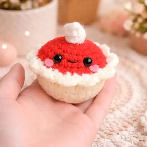 May include: A crocheted pie with a red top, white crust, and a small white pom-pom. The pie has black eyes and pink cheeks, giving it a friendly appearance. The pie is held in a hand.