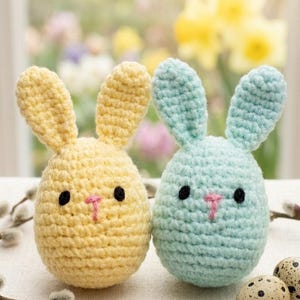 May include: Two handmade crochet Easter bunny decorations. One is yellow, the other is light blue. Both have black eyes and pink noses. The bunnies are egg-shaped with upright ears. Decorative quail eggs and pussy willow branches are also present.