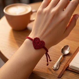 May include: A red crochet heart bracelet. The bracelet features a heart-shaped charm in the center, connected to a thin, adjustable band. The band has small gold-colored beads at the ends. The bracelet is worn on a wrist.