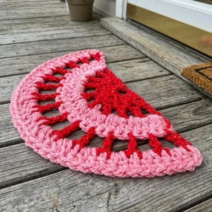 May include: A handmade, half-circle doormat in the shape of a watermelon slice. The mat is crocheted with pink and red yarn, with a textured design. The red yarn has a glittery effect. Placed on a wooden porch.