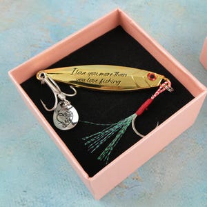 Könnte beinhalten: Ein goldfarbener Angelköder mit der Gravur "I love you more than you love fishing" wird in einer rosa Geschenkbox präsentiert. Der Köder hat einen silbernen Haken und einen Anhänger mit der Aufschrift "Bite Me". Ein einzigartiges Geschenk für Angler.