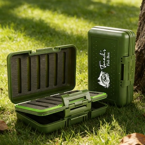 Könnte beinhalten: Zwei olivgrüne Angelkisten, eine offen und eine geschlossen, liegen auf Gras. Die offene Box zeigt einen dunklen Schaumstoffinnenraum mit Rillen. Die geschlossene Box hat den Text "Tenoch's Fish Box" und eine Fischgrafik.