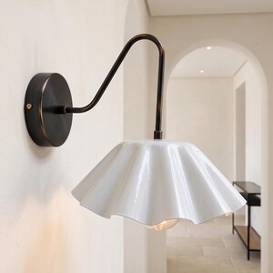 May include: A wall-mounted lamp featuring a dark bronze-toned metal arm and a circular wall plate. The fixture includes a glossy white, fluted, bell-shaped shade that directs light downwards, exposing a single light bulb at the centre.