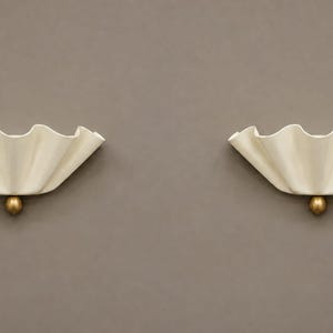 May include: A pair of wall lights featuring off-white, ruffled, shell-like shades with a scalloped edge. Each light is anchored at the bottom by a small, spherical, metallic gold-toned accent piece that connects the shade to the wall mount.