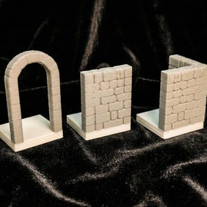 Può includere: Tre modelli architettonici in miniatura grigi con texture di pietra. Uno è un arco, e gli altri due sono sezioni di muro. Tutti sono montati su basi rettangolari bianche, adatti per giochi da tavolo o modellismo.